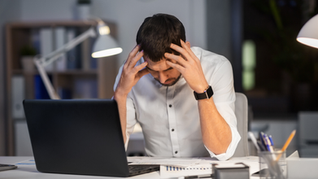Man in white shirt holds head in frustration at desk with laptop. Papers and pen holder nearby. Office setting, muted colors, tense mood.