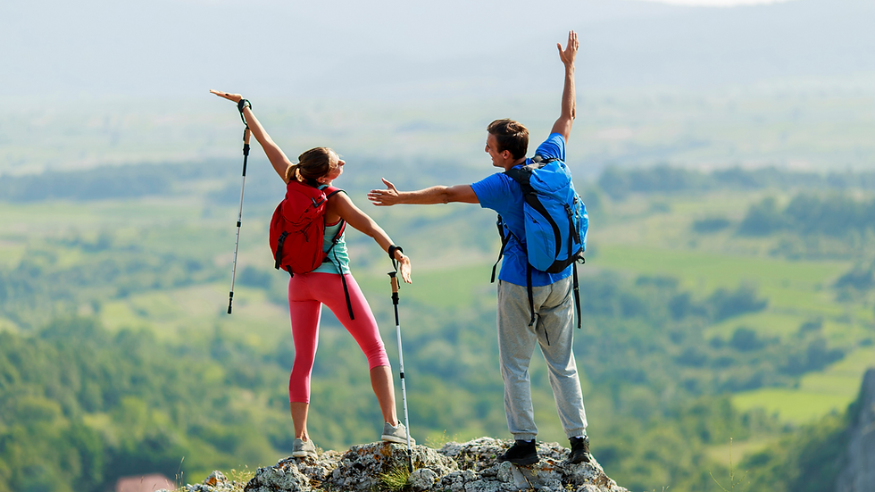 Two hikers with backpacks and poles celebrate atop a mountain, arms raised. Bright outfits against a lush, green valley backdrop.