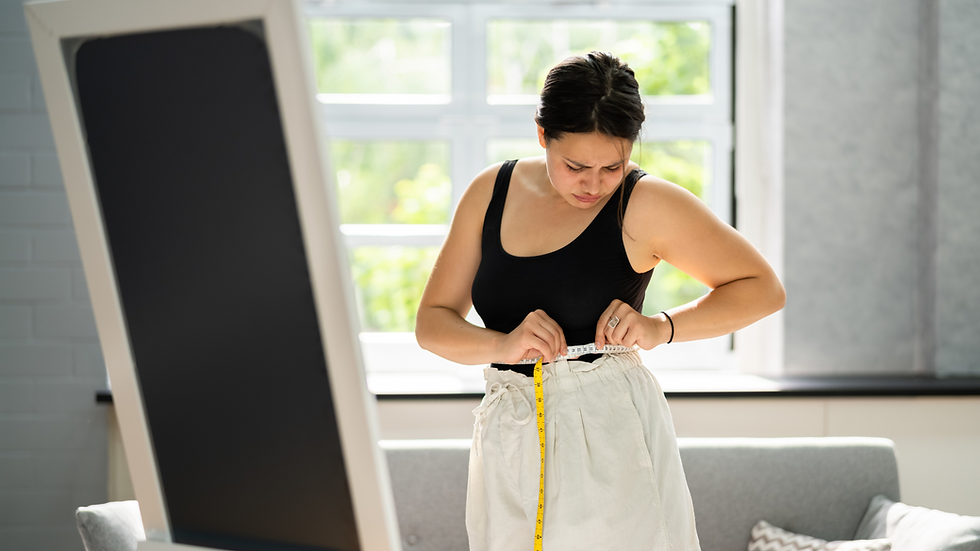 Woman measuring waist with tape in bright room, wearing black top and white skirt. A mirror and sofa are in the background.