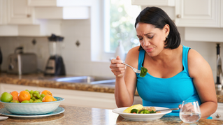 Woman in blue tank top frowns at broccoli on fork, seated in a bright kitchen. Plate of fruits and glass of water are on the counter.