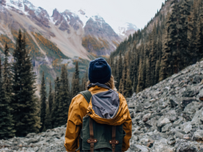 Person in yellow jacket and blue hat with backpack stands on rocky mountain trail, surrounded by pine trees, facing snowy peaks.