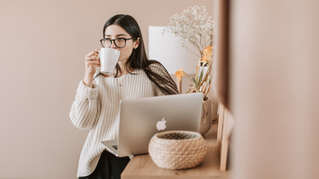 Woman in glasses sips from a white mug beside an open MacBook at a cozy beige desk with flowers.