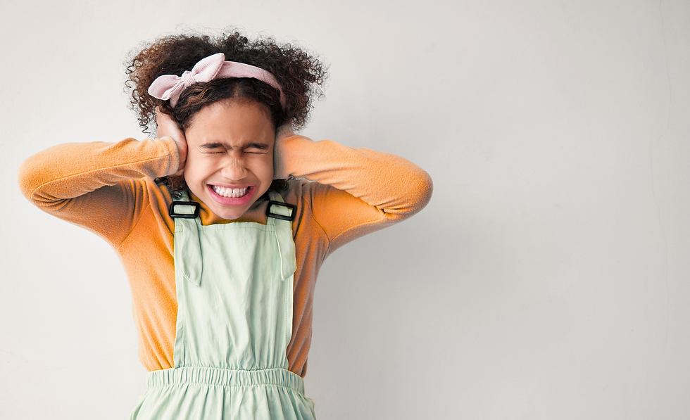 Child wearing orange sweater and green overalls presses hands over ears, eyes shut tight, with a pink headband. Plain white background.