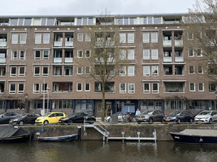 Gevel van een bakstenen gebouw met balkons en ramen. Voor het gebouw staan auto's geparkeerd. Op de voorgrond is een gracht met boten.