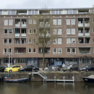 Gevel van een bakstenen gebouw met balkons en ramen. Voor het gebouw staan auto's geparkeerd. Op de voorgrond is een gracht met boten.