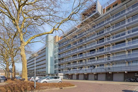 Moderne flat met lange balkons, geparkeerde auto’s en kale bomen op een zonnige dag.