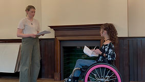 Leah and fellow Actress Anna performing a scene. They stare intenseley at eachother, both tearful. Leah is in her wheelchair, Anna stands opposite her. They both hold scripts in their hands, rehearsing.