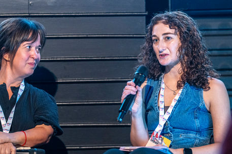Leah speaks into a microphone with a Sky Kids lanyard around her neck, wearing a blue denim waistcoat. Nichola is turned towards Leah, smiling at what she’s saying.