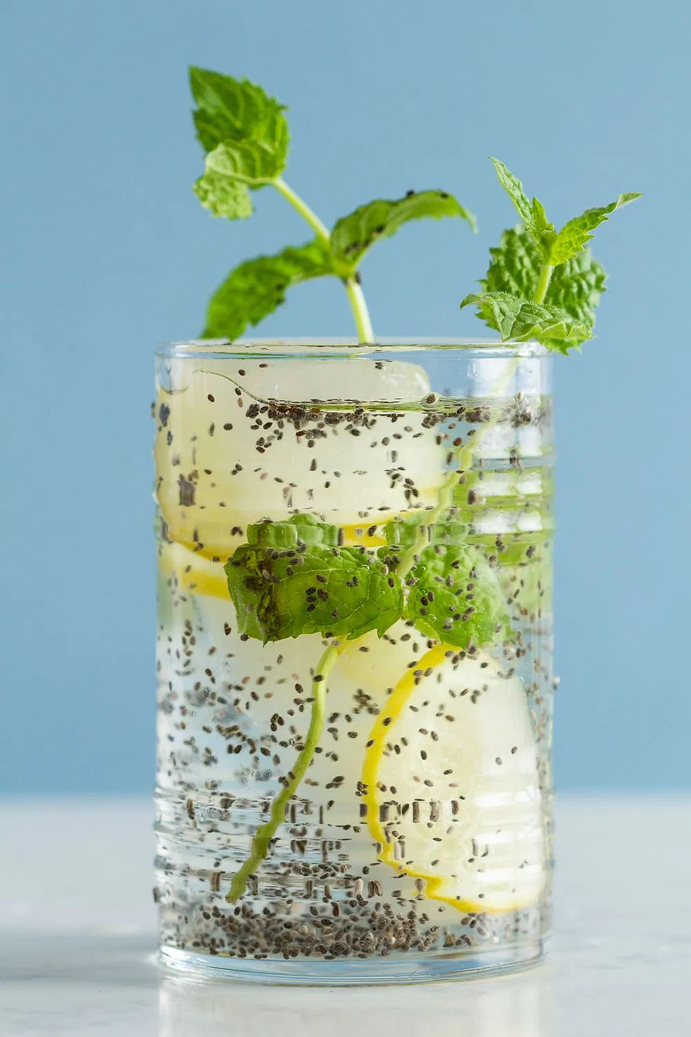 Chia seed drink with lemon slices, ice, and mint leaves in a glass. Blue background, refreshing and vibrant mood.