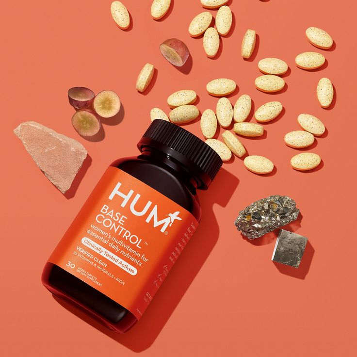 Orange HUM Base Control multivitamin bottle on a matching background, surrounded by yellow pills, rock, minerals, and grape slices.