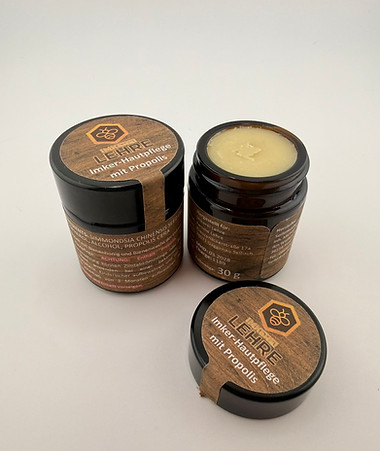 Nahaufnahme eines geöffneten braunen Glastiegels mit naturfarbener Propolis-Salbe sowie zwei geschlossenen Dosen der „Imker Hautpflege mit Propolis“ auf hellem Hintergrund. Die handgefertigte Naturkosmetik aus der Imkerei ist in kleinen 30-g-Tiegeln mit Holzoptik-Etikett verpackt und zeigt die cremige Konsistenz der pflegenden Hautsalbe aus Bienenwachs und Propolis.