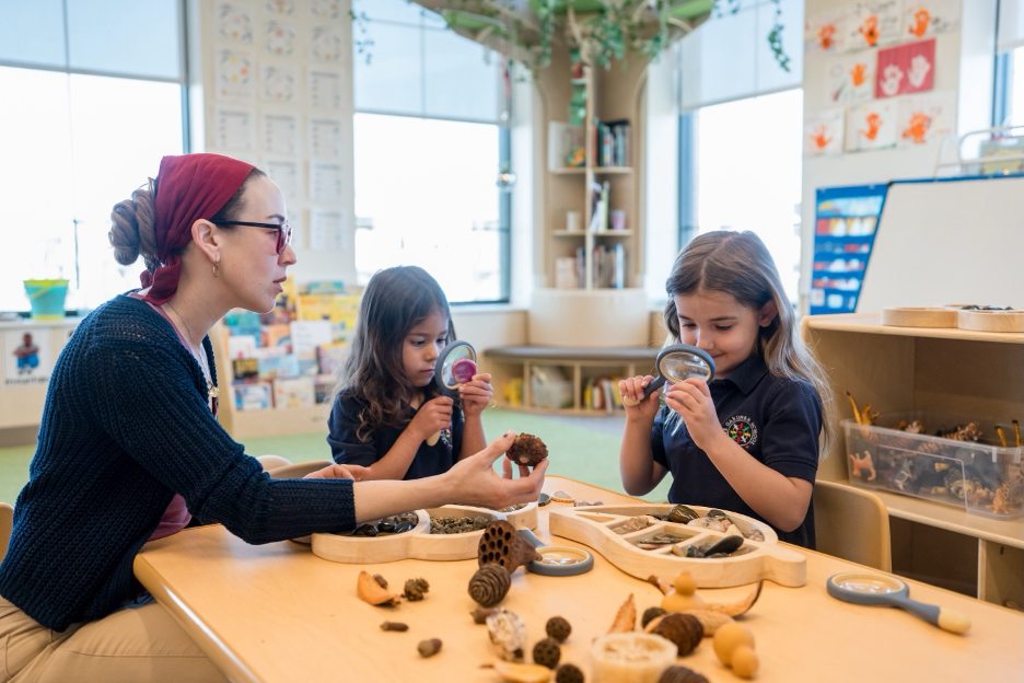 Teacher and two preschool girls exploring pinecones and nature items with magnifying glasses