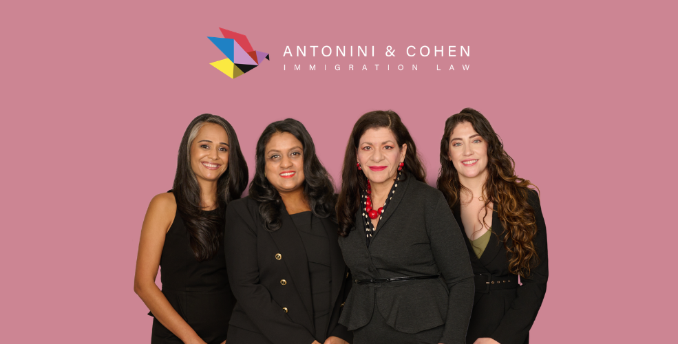 Immigration Attorneys Sonal Shah, Nisha Karnani, Carolina Antonini, and Kathleen Hoyos in black suits smile against a pink background beneath the Antonini & Cohen Immigration Law logo.