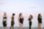 Five women of various body types and ethnicities stand on a beach in black athletic wear, spaced apart and facing different directions. The serene ocean and pastel sky create a calm backdrop, emphasizing diversity and individuality.