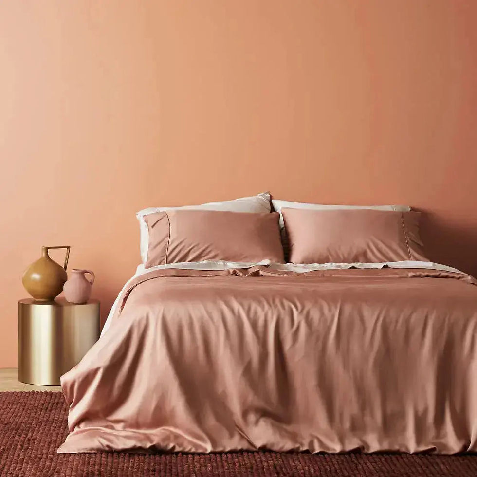 A cozy bedroom setup featuring a bed dressed in soft, blush-toned sheets and pillowcases against a warm terracotta-colored wall. A modern gold side table with minimalistic ceramic decor completes the look, creating a soothing and stylish space.
