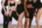 A group of women in various black athletic wear standing on a beach with a serene ocean background. The image highlights diverse body types and a sense of unity and confidence among the women.