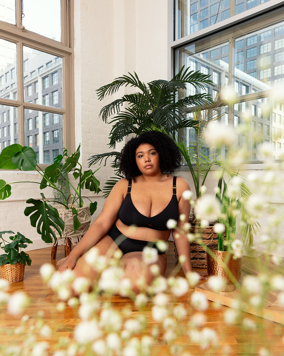 A model sits confidently on a wooden floor in a bright studio, wearing a black lingerie set and surrounded by lush green plants in woven and copper pots. Soft natural light flows through large windows, highlighting her relaxed pose and serene expression. The foreground features out-of-focus flowers, adding a gentle, layered effect to the image.