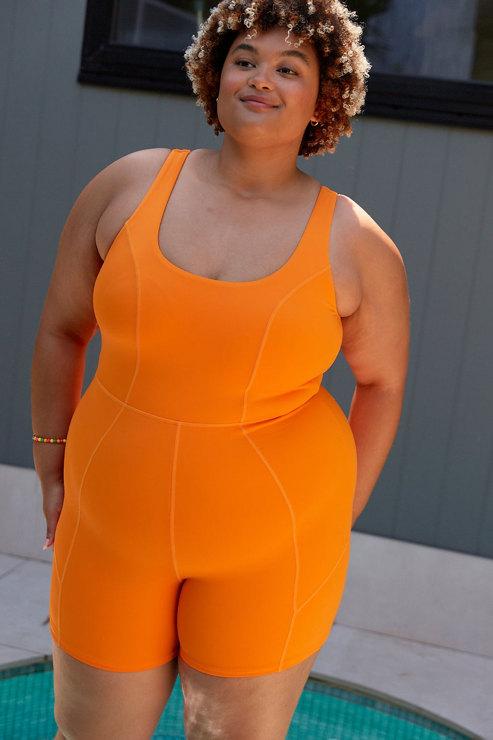 A woman confidently models a vibrant orange one-piece swimwear designed for versatility, suitable for the beach, Pilates, or brunch. The outfit highlights sleek stitching and a flattering fit, merging functionality and style for various occasions.