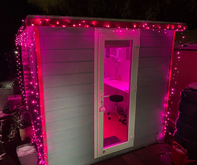 A compact, purpose-built pet grooming pod illuminated with pink LED lights. The modern structure features a white door with a glass panel, revealing a clean and organised grooming station inside. The exterior is decorated with string lights, creating a warm and inviting atmosphere in the snowy outdoor setting.