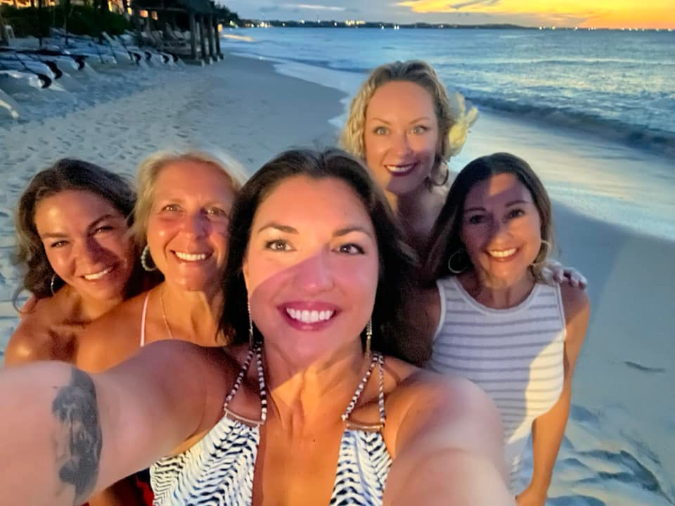 Five female friends pose for a smiling selfie on a beach