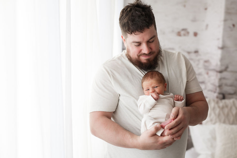 Dad cradles a newborn in a bright room with white walls. Both wear white. The scene is calm and intimate.