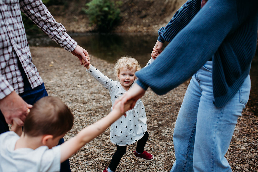 A girl in a polka-dot dress smiles, holding hands with adults and a child, forming a circle on a gravel path by a river. Casual, joyful scene.