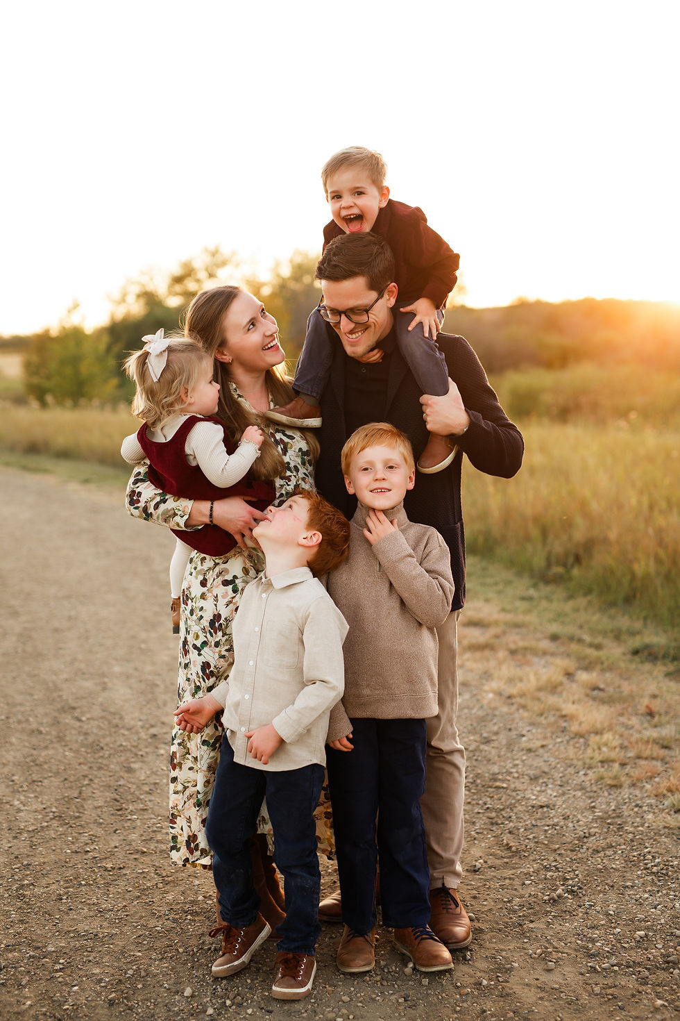 Happy family of six on a country road at sunset, children clustered around parents, one boy on dad’s shoulders smiling during edmonton fall family photography session