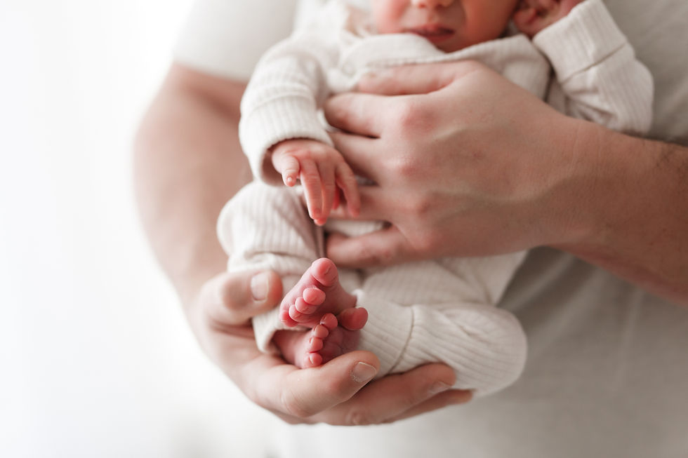 Adult gently holds a newborn in white clothes, highlighting tiny feet. Soft lighting creates a tender, serene atmosphere.