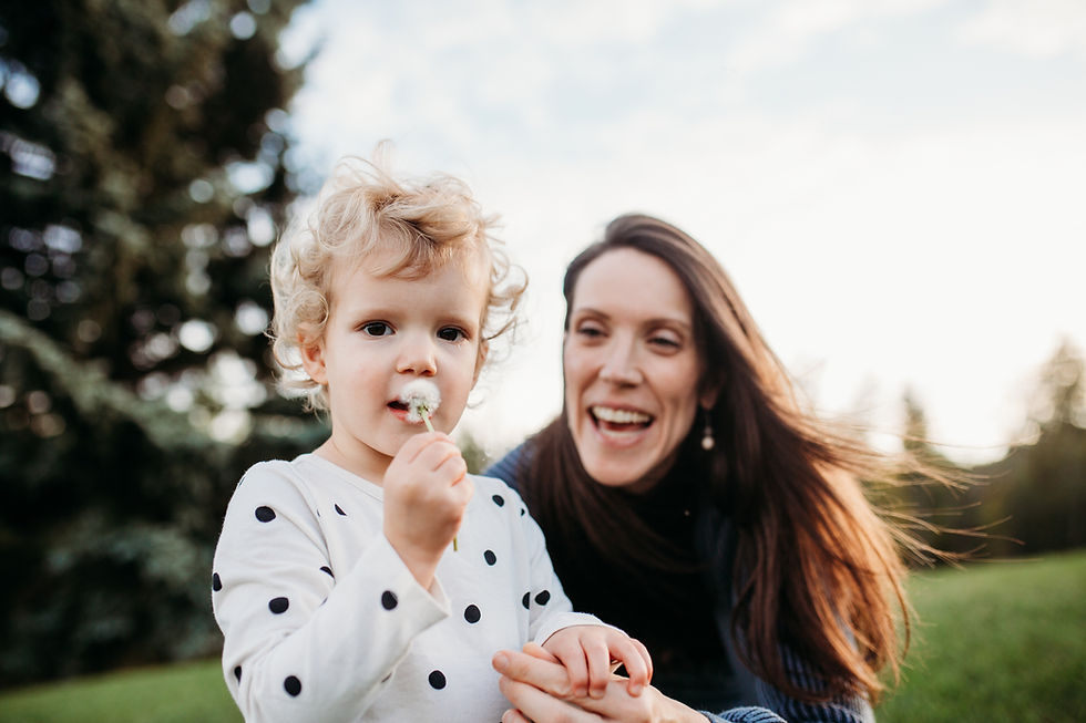 Child with curly hair holds a dandelion, wearing a polka dot shirt. Smiling Mom behind them. Green grass and trees in the background.