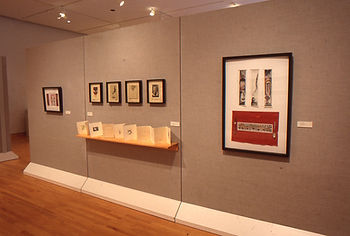 A mixed media studio art installation titled Objects of Devotion from an MFA thesis exhibition, displaying framed work and an accordian book in a gallery setting