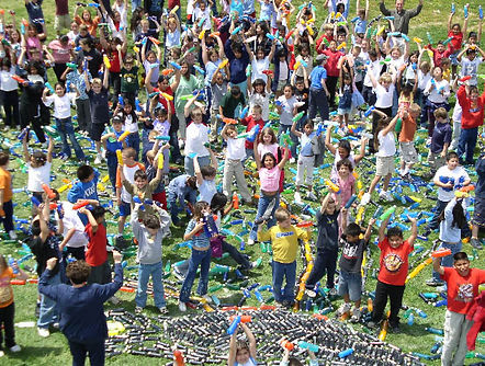 A large group of elementary school students gathered together on a grass field, actively participating in the collaborative construction of a large-scale interactive art installation.