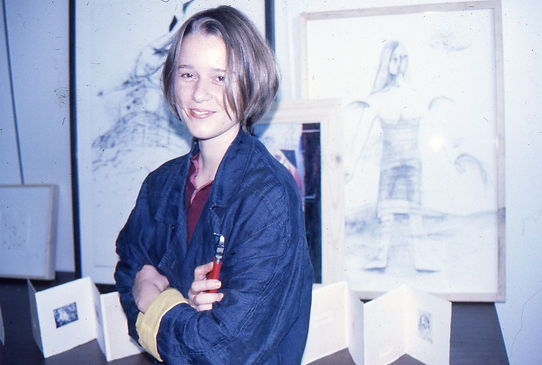 A front-facing portrait of a female artist-educator standing in a studio workspace alongside an original framed contemporary work on paper before a museum exhibition.