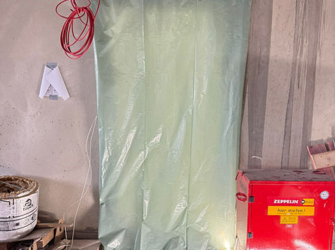 Grünes Material in Plastikverpackung, rotes Werkzeug daneben, auf grauer Oberfläche.