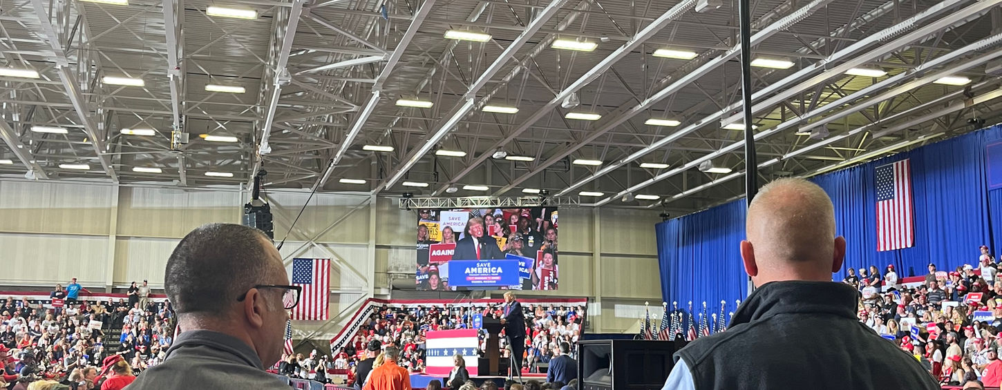 Two men watching a crowded event with a speaker on stage
