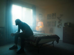 A man sitting on the edge of a bed in a dark room, head hanging, hands folded.