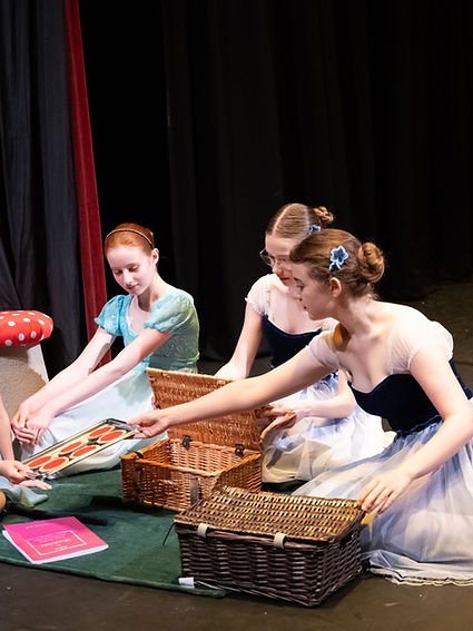 Three dancers in costumes sitting with a picnic basket on stage