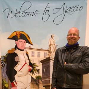 Smiling man with left hand inside his leather jacket, standing front of a poster drawing representing Napoleon with uniform and his hand inside his jacket. Text written on poster Welcome to Ajaccio