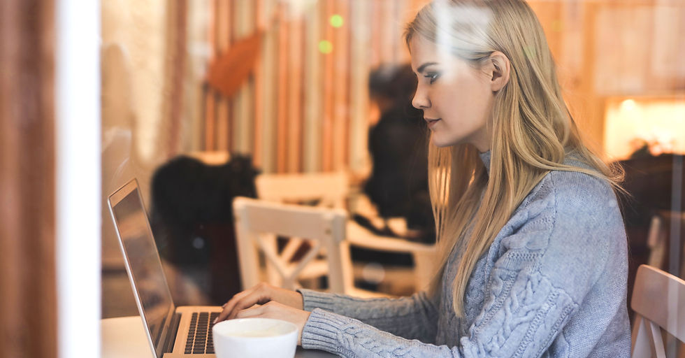 A focused woman works on her laptop at a sunlit cafe table, others visible in the warm blurred background
