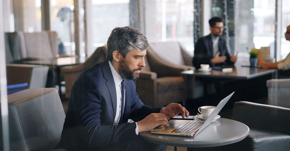A man works on a laptop in a cafe while another person sits nearby.
