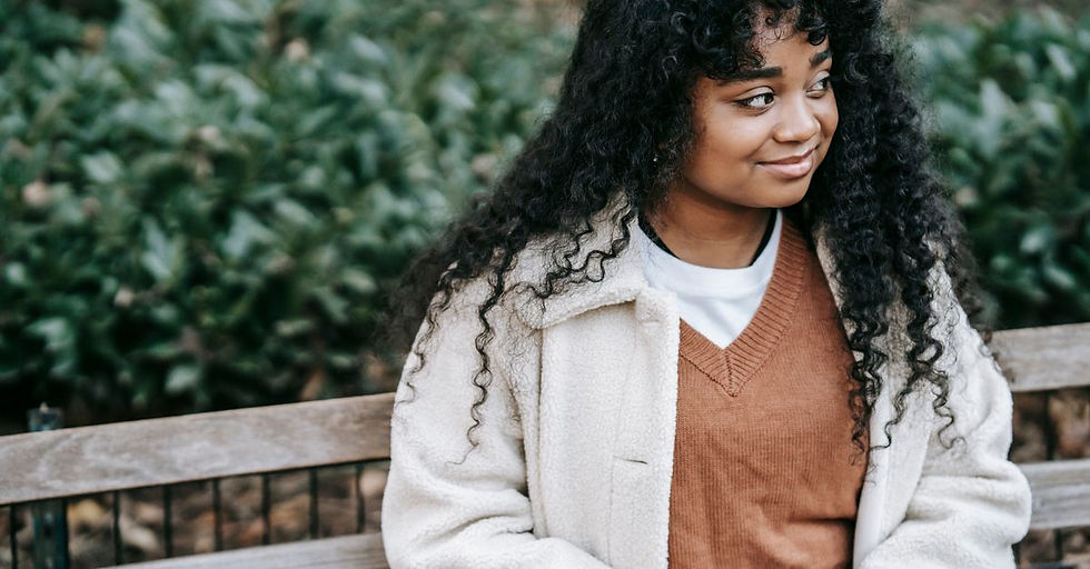 A young woman in a cream coat sits on a park bench, smiling softly as she looks to the side