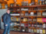 Man in black gear smiling in front of shelves filled with colorful liquor bottles. Bright orange walls and framed pictures in the background. AvVida.co.uk - Adventure Motorcycle Travel