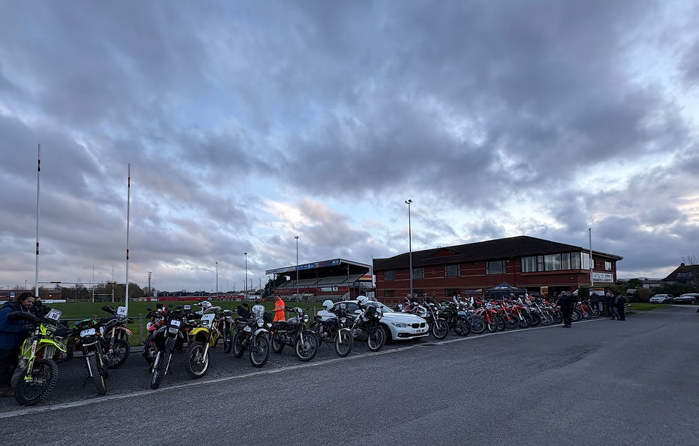 Motorcycles parked in a row outside a brick building under a cloudy sky. People stand nearby. A white car is at the end of the row. AvVida Micro Adventures - Life is an Adventure. La Vida es Una Aventura.