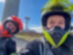 Two people in colorful helmets take a selfie near wind turbines. Bright blue sky and grassy landscape create an adventurous mood. AvVida.co.uk - Adventure Motorcycle Travel