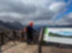 Person in orange helmet on wooden lookout deck, overlooking mountainous landscape. "Mirador de Piedrasluengas" sign visible. Cloudy sky. AvVida.co.uk - Adventure Motorcycle Travel
