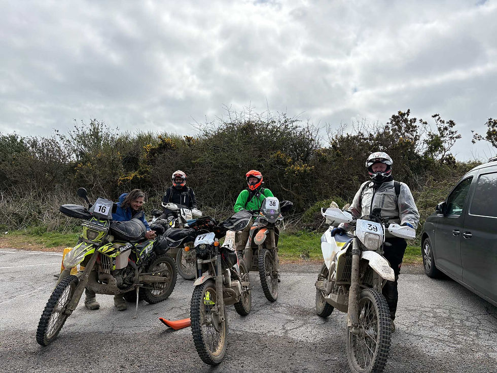 Four people with helmets and dirt bikes, each numbered, rest on a road with bushes and a cloudy sky in the background, near a parked van. AvVida Micro Adventures - Life is an Adventure. La Vida es Una Aventura.