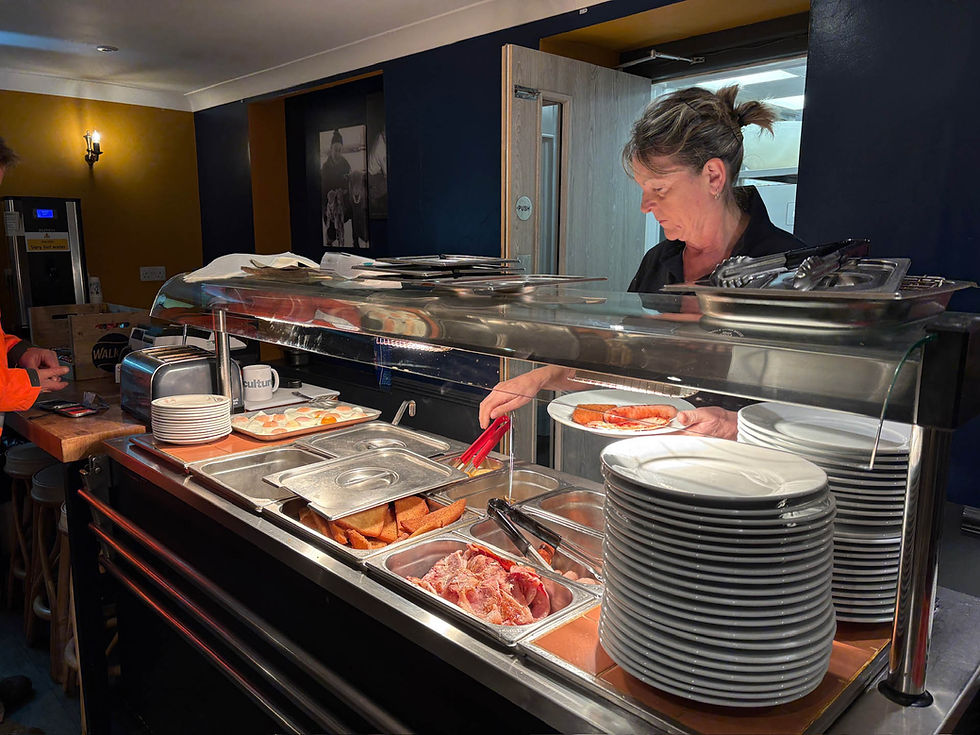Server preparing breakfast at a buffet with toasters and food trays. Plates stacked nearby. Warm lighting creates a cozy atmosphere. AvVida Micro Adventures - Life is an Adventure. La Vida es Una Aventura.