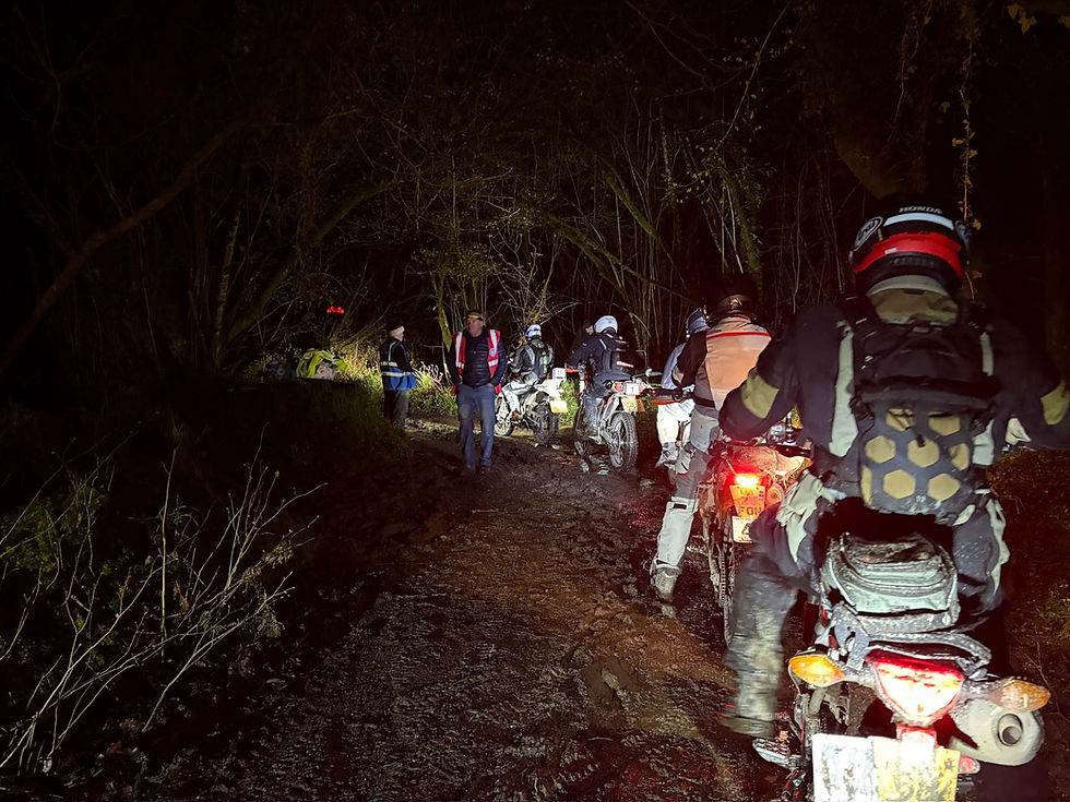 Motorcyclists ride through a muddy forest path at night. Bright headlights illuminate the damp surroundings. Observers in safety vests watch. AvVida Micro Adventures - Life is an Adventure. La Vida es Una Aventura.