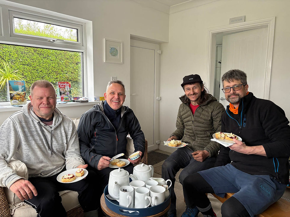 Four men sit indoors enjoying scones with cream and jam. White teapots and cups are on a table. The mood is cheerful and relaxed. AvVida Micro Adventures - Life is an Adventure. La Vida es Una Aventura.
