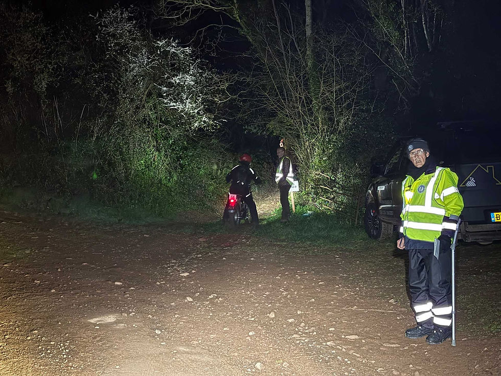 Person in high-visibility gear stands beside a parked vehicle at night. Another person on a motorbike heads into a forest path, with flashlights illuminating the scene. AvVida Micro Adventures - Life is an Adventure. La Vida es Una Aventura.