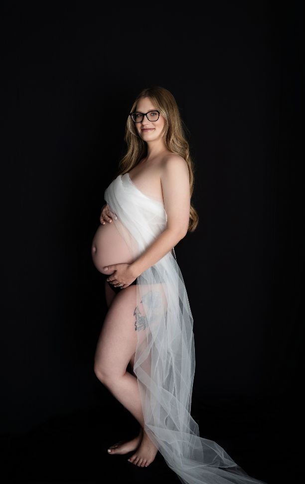 Pregnant woman in sheer fabric posing in profile against a dark background during a maternity boudoir shoot